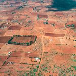 Somewhere in the land of the Masai, Kenya - March 24th 2022. Aerial view of the red soiled savannah speckled with bushes in Kenya during the drought of 2022. Small traditional Masai homesteads with grain fields can be seen all over the beautiful landscape.